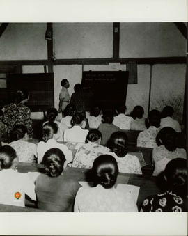 Seorang wanita maju di depan kelas sedang belajar menulis didampingi oleh seorang guru.