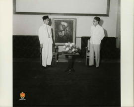 Dua orang pejabat sedang bercakap-cakap dengan latar belakang gambar Pangeran Diponegoro.